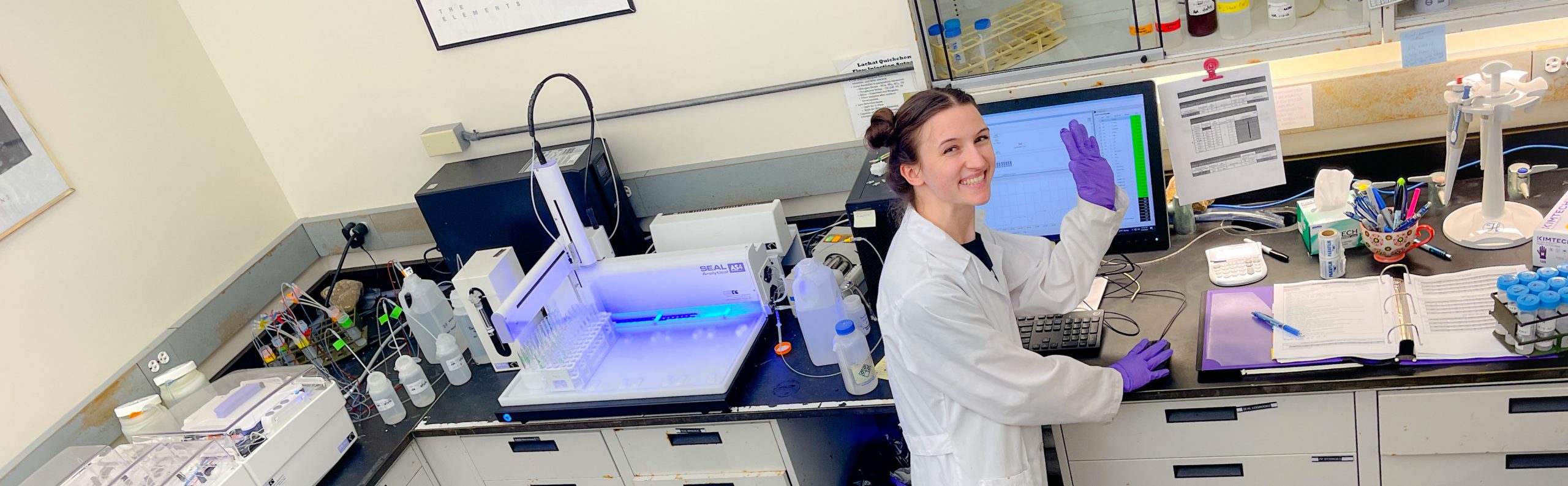 A scientist in a white coat looks away from her lab computer to smile and wave at the camera