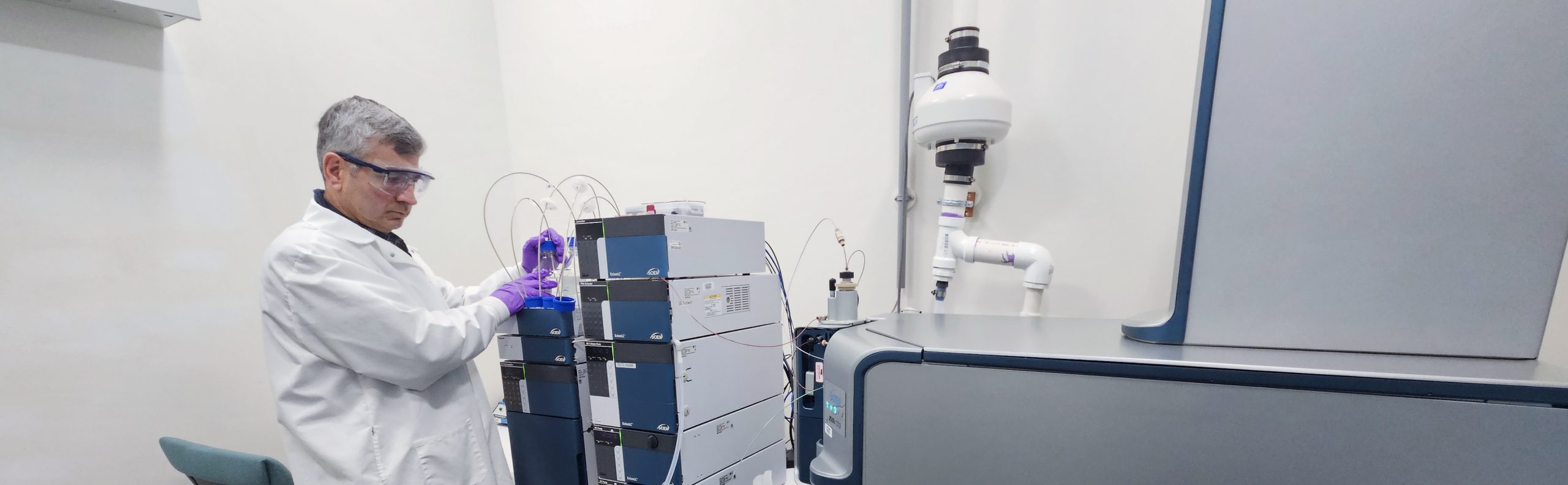 A researcher in a white coat, safety glasses, and purple gloves works with large machinery in a lab